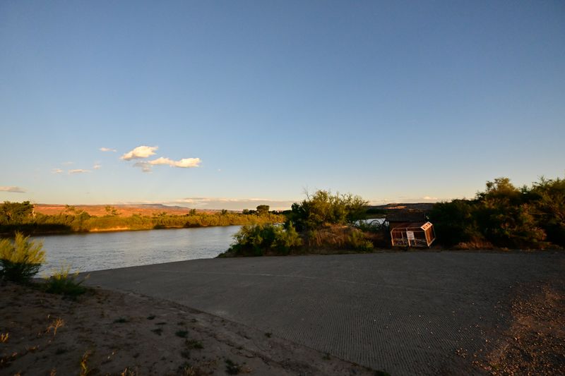 Concrete boat ramp sloping into the calm Colorado River at the Cisco takeout, evening light warming the far bank and a small trash enclosure among the tamarisk.