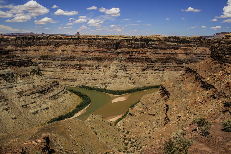 View from the canyon rim of the confluence of the Green and Colorado rivers, two muddy channels meeting around a pale sandbar far below stratified canyon walls under scattered clouds.