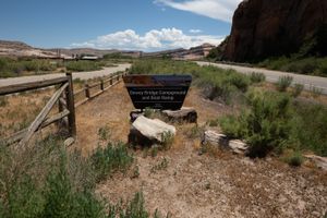 BLM "Dewey Bridge Campground and Boat Ramp" sign in sage beside River Road, a red cliff rising at right.