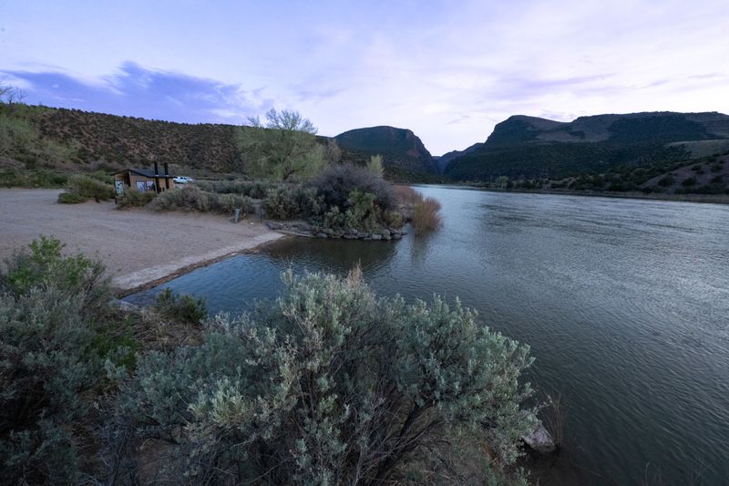 Green River sliding past the Gates of Lodore put-in at dusk, with a gravel launch area, information kiosk and outbuilding on the left and the dark canyon gates opening downstream.