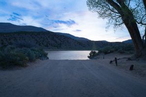 Dirt boat-ramp lane running down to the Green River at dusk at the Gates of Lodore put-in, a large cottonwood on the right and sagebrush flanking both sides under a pink-streaked sky.