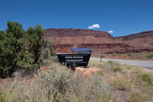 BLM "Hittle Bottom Campground" sign on a sandstone base beside River Road, juniper at left and red cliffs behind.