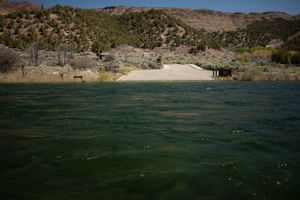Paved Little Hole boat ramp seen head-on from the water, dark green rippled river in the foreground and juniper-dotted hills rising behind under a clear midday sky.