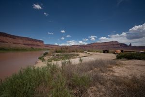 Wide gravel lot beside the Colorado at Lower Onion Creek, an outfitter bus and stacked orange rafts at the far end.