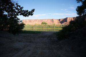 Sandy track running down to the Green River at Mineral Bottom, sunlit rimrock across the water above a green bank.