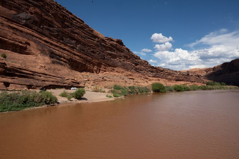 The muddy Colorado below the Moab bridge ramp, with the launch flat and parked vehicles under a long tilted red wall.