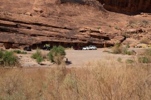 Gravel staging lot under an overhanging red wall at the Moab bridge ramp, parked vehicles and a cyclist crossing the flat.