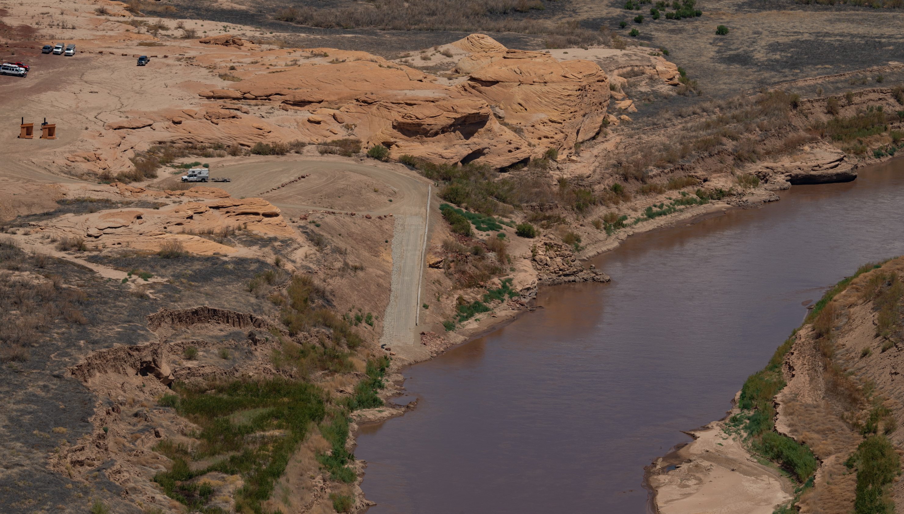 Elevated view of the North Wash take-out: a single-lane ramp running straight down a bare dirt bank into the muddy Colorado River, with a graded dirt turnaround above it, a truck and boat trailer parked on the flat, and more vehicles and two vault toilets on the slickrock bench beyond.