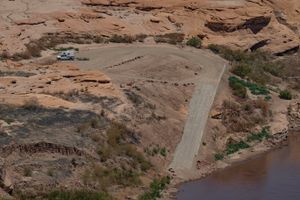 The North Wash ramp seen from above — a narrow paved lane with a low concrete barrier along its right edge and loose riprap along its left, dropping down a steep dirt bank to brown water. A single truck with a boat trailer is parked on the rock-lined dirt turnaround at the top.