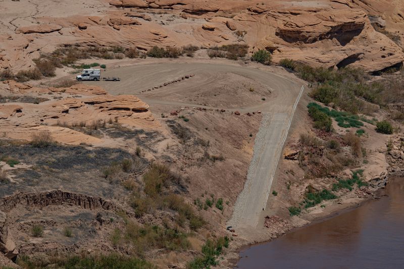 The North Wash ramp seen from above — a narrow paved lane with a low concrete barrier along its right edge and loose riprap along its left, dropping down a steep dirt bank to brown water. A single truck with a boat trailer is parked on the rock-lined dirt turnaround at the top.