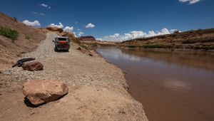 Ground-level view at the foot of the North Wash ramp, where the paved surface meets a cobble and sand shoreline beside the brown Colorado River. A flatbed camper truck with a mountain bike on the back sits partway up the ramp; sandstone boulders and coiled black drainage pipe lie on the bank, with canyon walls and cumulus cloud downstream.