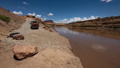 Ground-level view at the foot of the North Wash ramp, where the paved surface meets a cobble and sand shoreline beside the brown Colorado River. A flatbed camper truck with a mountain bike on the back sits partway up the ramp; sandstone boulders and coiled black drainage pipe lie on the bank, with canyon walls and cumulus cloud downstream.