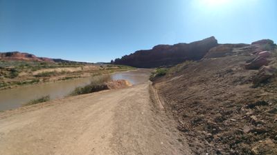 Dirt access road descending to the silt-laden Colorado River at the North Wash take-out, dark red cliff bands downstream under a harsh midday sun.