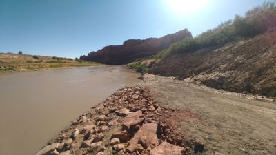 Broken red rock and cobble shoreline sloping into the muddy Colorado River at the North Wash take-out, tamarisk-covered bank rising to the right and dark cliffs downstream.