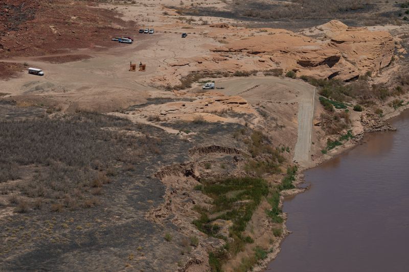 Elevated view of the North Wash staging area: a broad dirt flat holding a white shuttle bus, a trailer loaded with rafts, several parked vehicles and two vault toilets, with the graded turnaround and the ramp descending to the Colorado River on the right and an eroded sediment bank below.