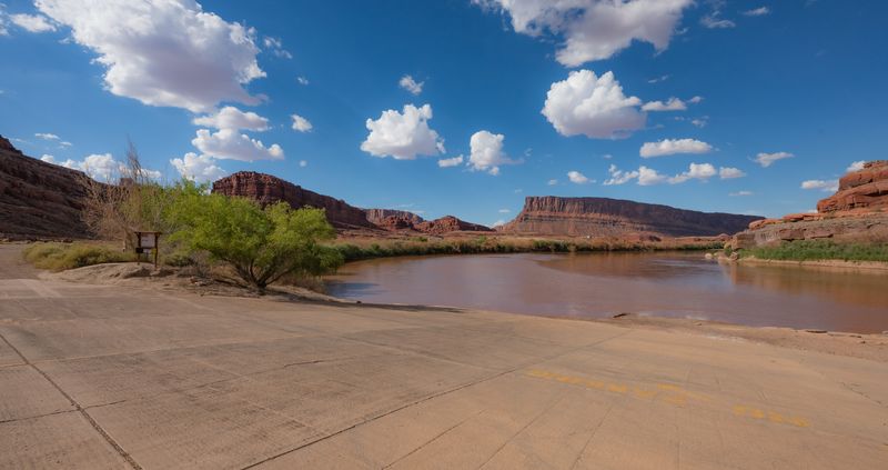 Wide concrete ramp meeting the brown Colorado at Potash, an information kiosk at left and red mesas under cumulus beyond.