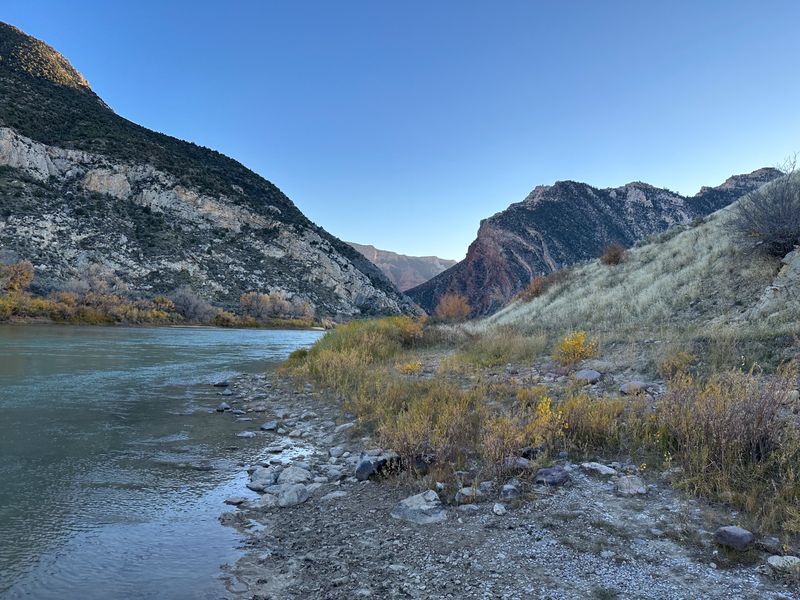 Cobble shoreline of the Green River at Rainbow Park, looking downstream toward the red tilted rock at the entrance of Split Mountain Canyon, with fall-yellow brush along the bank under a clear sky.