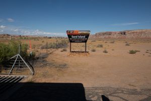BLM "Sand Island Boat Launch and Campground" sign beside the entrance cattle guard, mesas on the skyline behind.