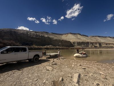 A pickup with an empty raft trailer backed to the water's edge at low water, a loaded gear raft floating in the shallows below badlands cliffs.