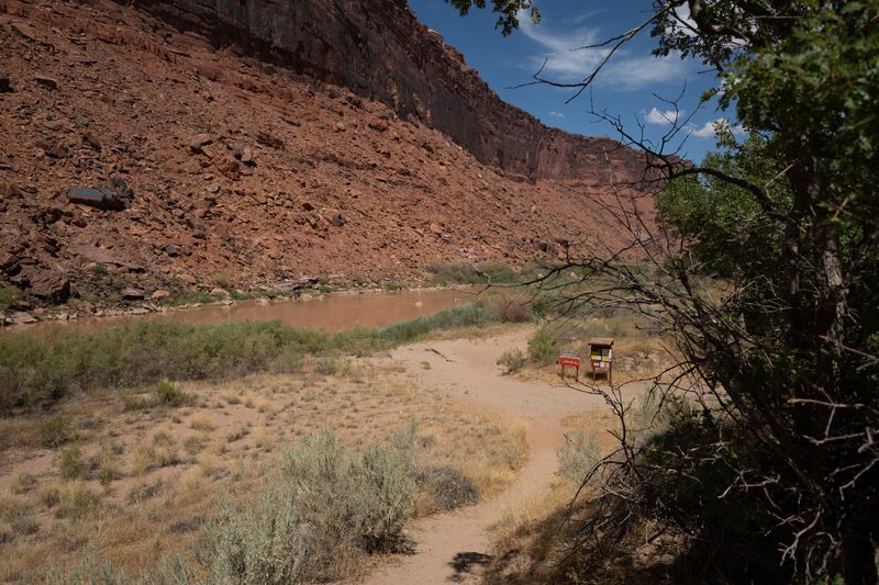 Sand track through sage down to the Colorado at Sandy Beach, a talus slope and dark cliff band rising across the water.