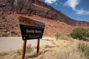 BLM "Sandy Beach" sign beside the paved lot, a camping marker post behind and a tall red cliff face above.