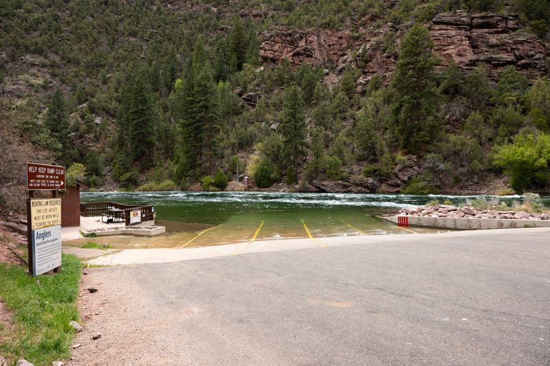 Concrete boat ramp with yellow lane stripes running into the green water of the Green River at the Spillway launch, with ramp-rules and angler-regulation signs at left and a pine-covered red rock canyon slope across the river.