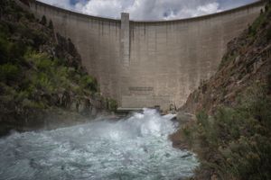 Water roars out at the base of Flaming Gorge Dam's curved concrete wall into a churning green-and-white river, between steep brushy canyon walls under a cloudy sky.