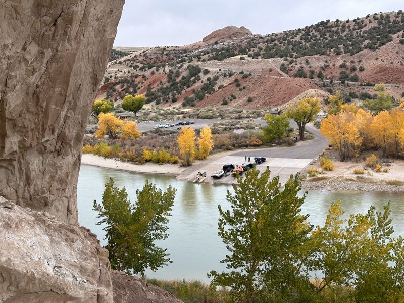 View from a sandstone ledge above the Split Mountain boat ramp on the Green River, with rafts and vehicles staged on the concrete ramp, cottonwoods in yellow fall color, red desert hills behind, and an overcast sky.