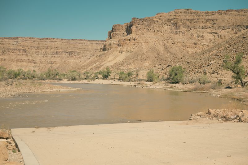 Wide concrete apron of the Swaseys boat ramp meeting the muddy Green River, with a band of green cottonwoods and brush across the water below tan desert cliffs under a clear sky.