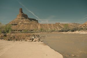 A spire-capped tan butte rises above the muddy Green River beside the concrete edge of the Swaseys boat ramp, with a bouldery shoreline, sparse spring greenery, and contrails crossing the blue sky.