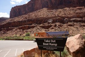 BLM "Take Out Boat Ramp" sign on a sandstone base beside the paved lot, red cliffs and talus rising behind.