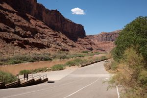 Paved staging lot with lane arrows and guardrail at Take Out Beach, the Colorado at left and the canyon opening downstream.