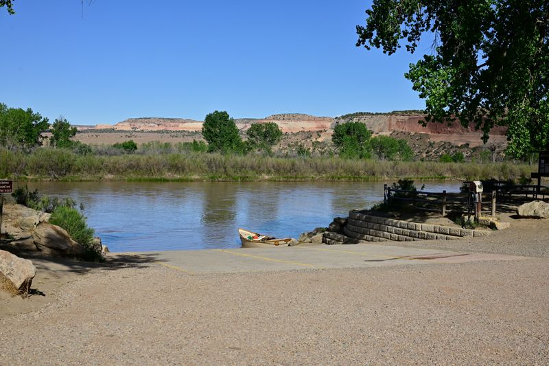 A loaded dory floats at the base of the striped concrete boat ramp at Westwater on the calm Colorado River, framed by a large cottonwood, with banded red and white sandstone cliffs across the water under a clear blue sky.