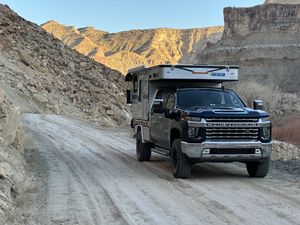 Dark pickup truck with a white pop-up camper driving a dusty dirt road cut along a rocky slope, golden-lit desert cliffs rising across the canyon.
