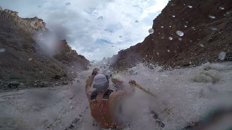Boat-mounted camera view of a rower in a helmet and life jacket pulling on the oars as a wall of brown whitewater explodes over the dory's bow, water beading the lens and dark canyon walls narrowing ahead.