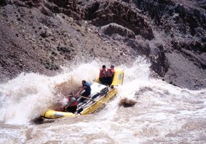 A yellow oar-rigged gear raft climbs a breaking silty wave in Big Drop 1 — the rower stands at the oars while passengers in orange life jackets brace at the bow and stern.