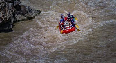 A red paddle raft with eight helmeted paddlers drives through churning brown whitewater beside dark, water-polished rock