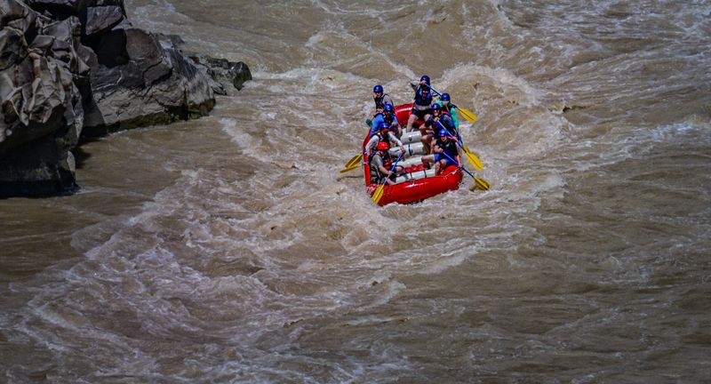 A red paddle raft with eight helmeted paddlers drives through churning brown whitewater beside dark, water-polished rock, yellow paddle blades raised mid-stroke.