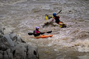 A kayaker in a purple helmet in an orange hardshell kayak and a paddler in a blue helmet in a yellow inflatable kayak work through churning brown whitewater beside dark gray boulders.
