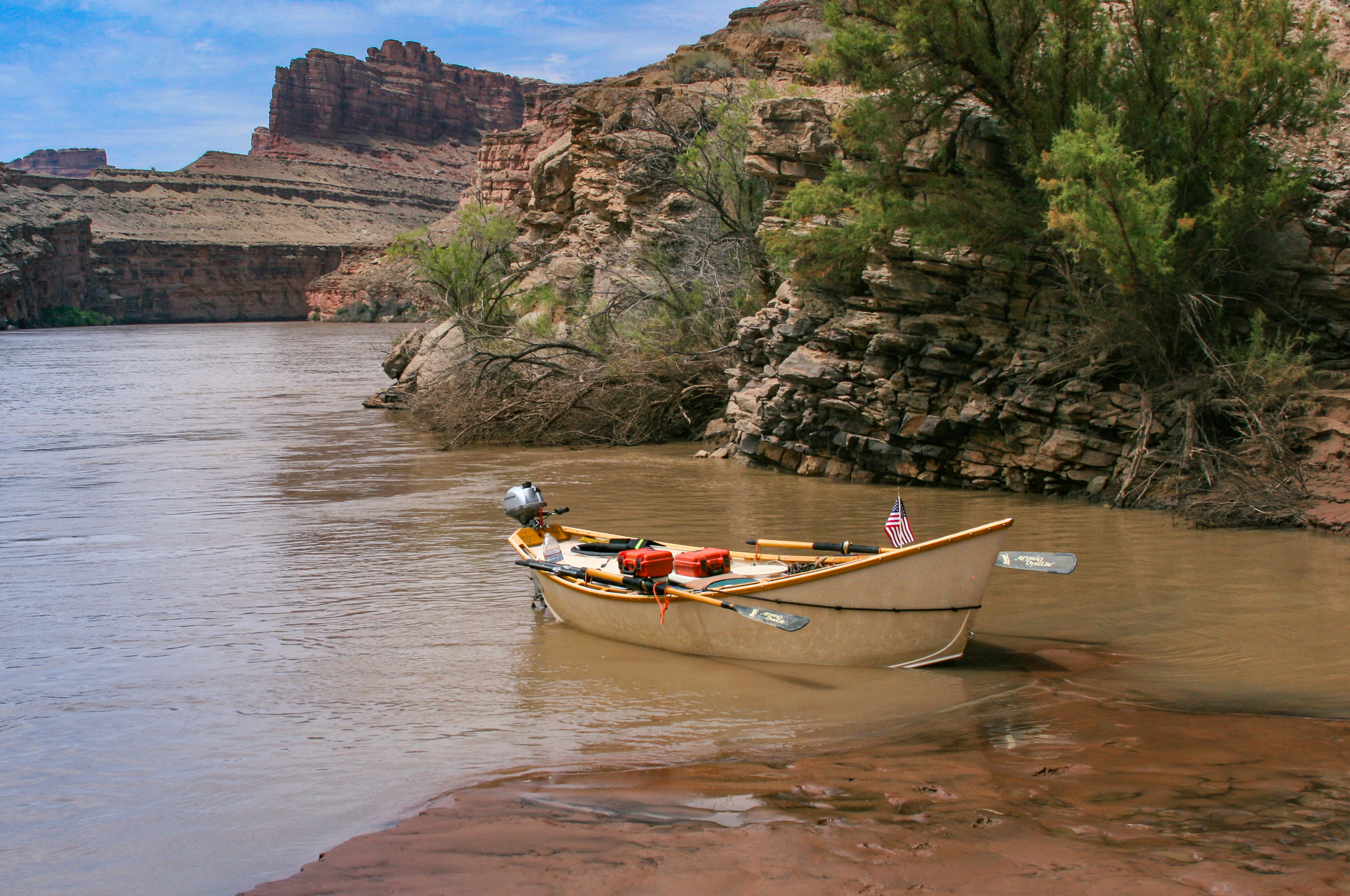 A wooden dory beached on a silty sandbar with a small outboard motor clamped to its transom, oars shipped and a red fuel can aboard