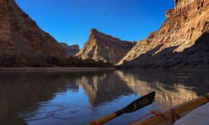 From the dory with an oar blade in frame, low sun lights a sandstone butte standing above a wide, mirror-still reach of river.