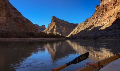 From the dory with an oar blade in frame, low sun lights a sandstone butte standing above a wide, mirror-still reach of river.