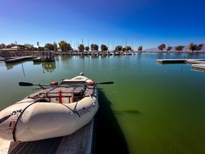 An eighteen-foot raft rigged with a rowing frame and a small outboard, tied alongside a dock in a green-water marina with sailboat masts and low hills beyond.