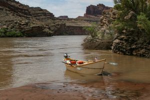 The same dory and clamped-on outboard seen from further back along the sandbar, tamarisk on the bank and layered red cliffs rising behind.