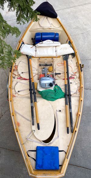 Overhead view into a wooden dory on dry land, a small outboard motor sitting in the footwell beside stowed oars, hatch covers and a green mesh bag.