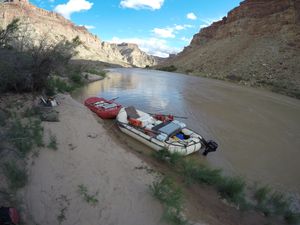 Two rafts tied along a sand bank — a red boat upstream, a white motor rig with an outboard on its transom downstream — with the silt-brown river curving away between gray and red canyon walls.