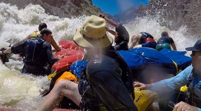A crew in life jackets and a straw hat braces on a double-rigged raft as a wall of whitewater breaks over the boat