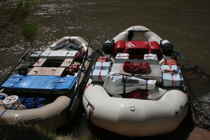 Two fully rigged gear rafts moored side by side on a muddy riverbank, seen from above, loaded with coolers, dry boxes, water jugs, dry bags and a rolled sleeping pad strapped to their frames.