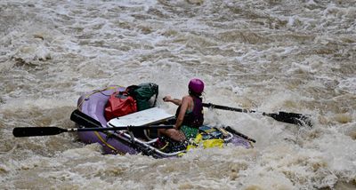 A rower in a purple helmet and life jacket leans into the oars of a small purple gear raft, rigged with a cooler