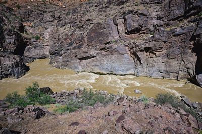 Muddy brown whitewater churns along the base of a sheer, dark rock wall at Skull Rapid in Westwater Canyon, seen from a rocky slope above a boulder-lined bank.